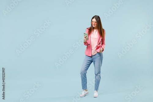 Full length young fun smiling happy woman 20s in pastel pink clothes glasses using mobile cell phone chatting online isolated on blue background studio portrait. People lifestyle technology concept