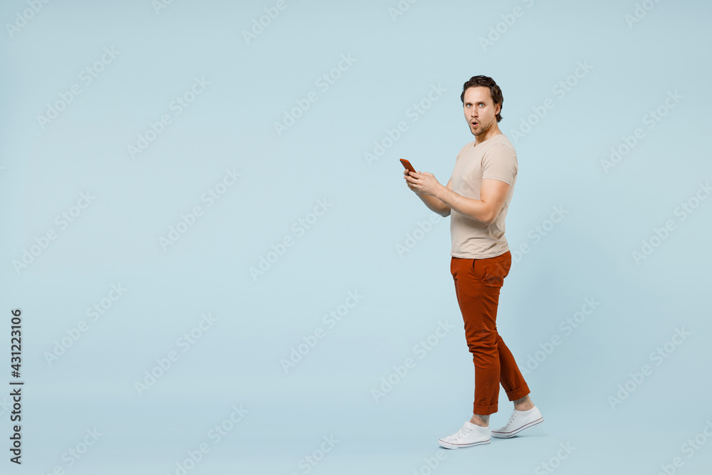 Obraz premium Full length side view young indignant confused man 20s wear casual basic beige t-shirt hold mobile cell phone look camera isolated on pastel blue background studio portrait. People lifestyle concept.