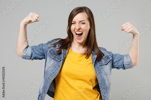 Young strong sporty fitness caucasian woman wearing casual denim jacket yellow t-shirt showing biceps muscles on hand demonstrating strength power isolated on grey color background studio portrait