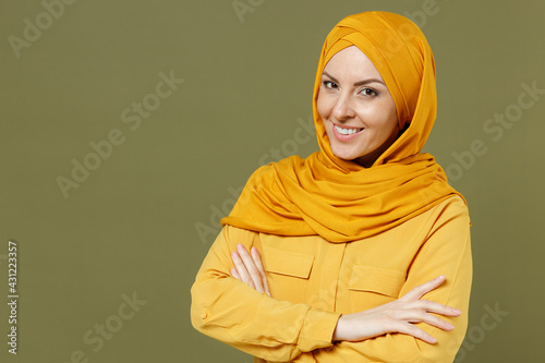 Smiling fun young arabian asian muslim woman in abaya hijab yellow clothes hold hands crossed folded isolated on olive green khaki color background. People uae middle eastern islam religious concept. © ViDi Studio