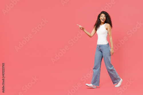 Full length young fun smiling happy friendly positive african american woman 20s wear casual white tank shirt walk go point index finger aside on copy space area mock up isolated on pink background