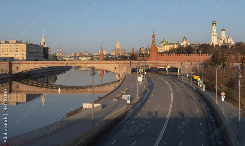 Fototapeta premium Early morning on the Moscow river. View of the Moscow Kremlin and the stone bridge. Reflection in water. Empty road along the embankment and a temple in the background