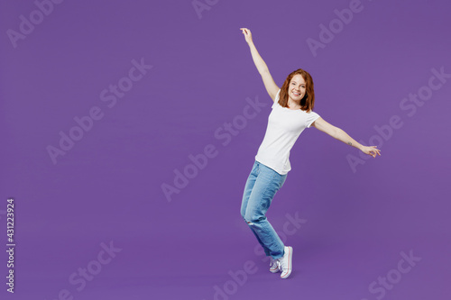Full length young happy fun cheerful redhead caucasian woman 20s wear white basic casual t-shirt jeans leaning back standing on toes dancing isolated on dark violet color background studio portrait