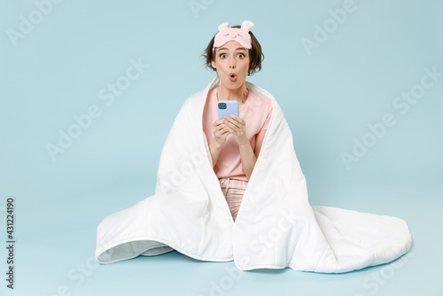 Young excited shock woman in pajamas jam sleep eye mask rest at home sit wrap cover under blanket duvet use mobile cell phone chat isolated on pastel blue background Good mood night bedtime concept