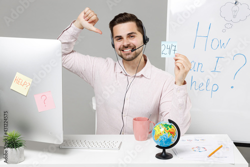 Young noctidial assistance employee business man in shirt sit work at office desk pc computer set microphone headset helpline hold 24 7 symbol card point finger on himself isolated on grey background photo