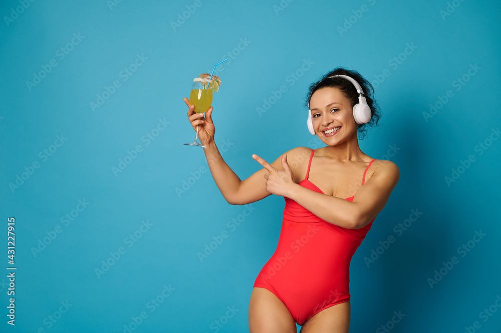 Attractive woman with a beautiful smile in a red swimsuit and headphones, standing on a blue background and pointing with a finger at a cocktail in her hand Attractive woman with a beautiful smile in a red swimsuit and headphones, standing on a blue background and pointing with a finger at a cocktail in her hand