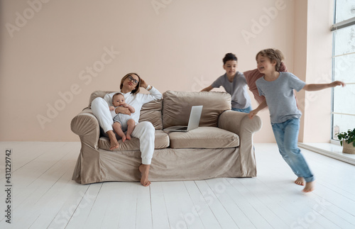 Young single mother working on laptop in loft sitting on couch while her sons running around her and shouting. High quality photo