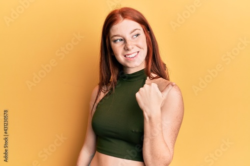 Young red head girl wearing casual clothes pointing thumb up to the side smiling happy with open mouth