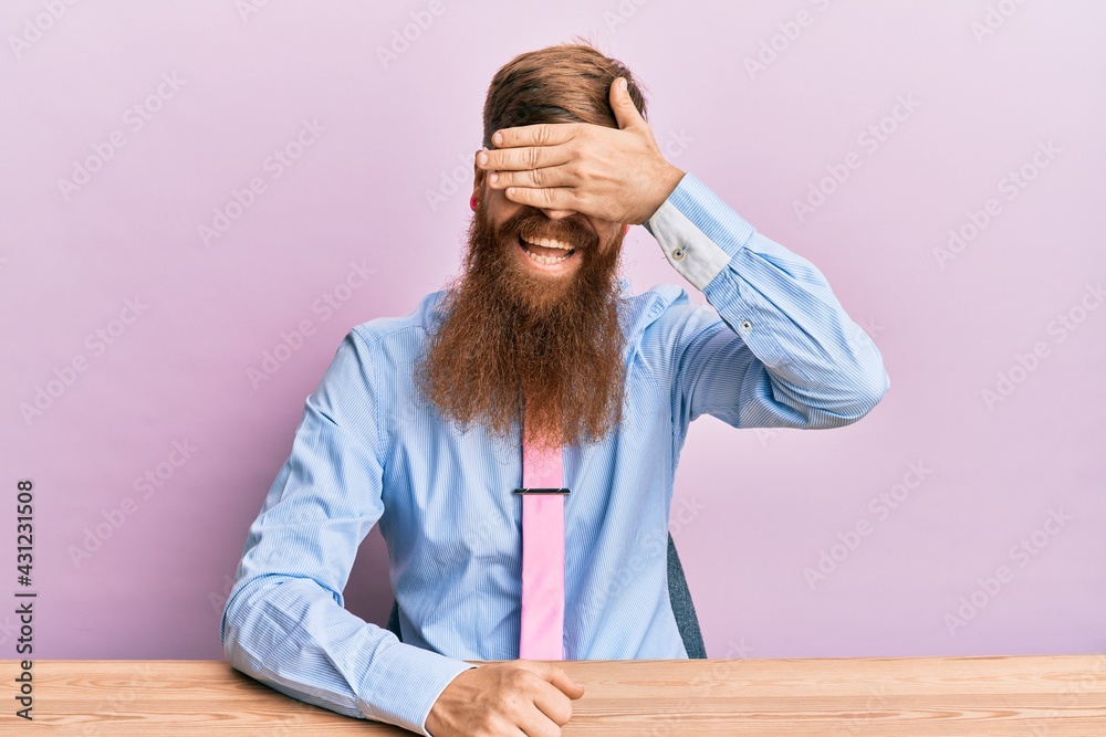 Young irish redhead man wearing business shirt and tie sitting on the table smiling and laughing with hand on face covering eyes for surprise. blind concept. Young irish redhead man wearing business shirt and tie sitting on the table smiling and laughing with hand on face covering eyes for surprise. blind concept.