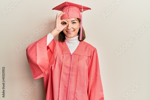 Young caucasian woman wearing graduation cap and ceremony robe doing ok gesture with hand smiling, eye looking through fingers with happy face.