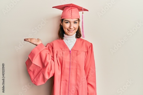 Young caucasian woman wearing graduation cap and ceremony robe smiling cheerful presenting and pointing with palm of hand looking at the camera.