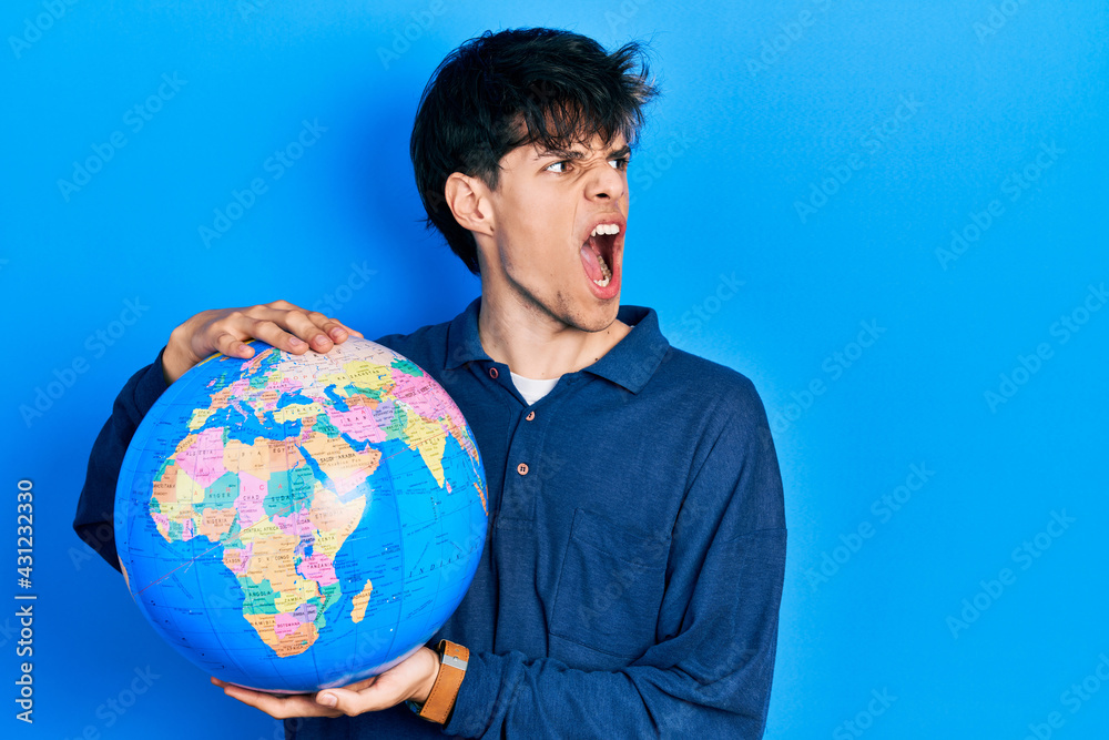 Handsome hipster young man holding world ball angry and mad screaming frustrated and furious, shouting with anger. rage and aggressive concept. Handsome hipster young man holding world ball angry and mad screaming frustrated and furious, shouting with anger. rage and aggressive concept.