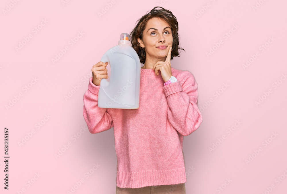 Young hispanic woman doing laundry holding detergent bottle serious face thinking about question with hand on chin, thoughtful about confusing idea Young hispanic woman doing laundry holding detergent bottle serious face thinking about question with hand on chin, thoughtful about confusing idea
