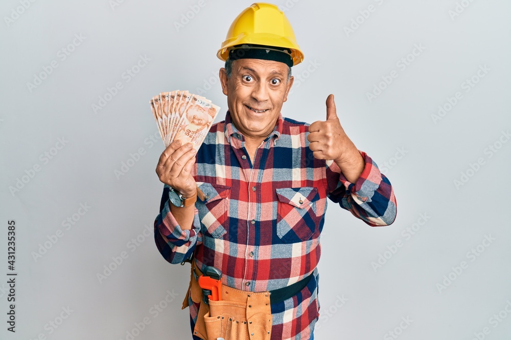 Senior hispanic man wearing handyman uniform holding turkish liras smiling happy and positive, thumb up doing excellent and approval sign
