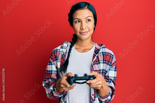 Beautiful hispanic woman playing video game holding controller smiling looking to the side and staring away thinking.