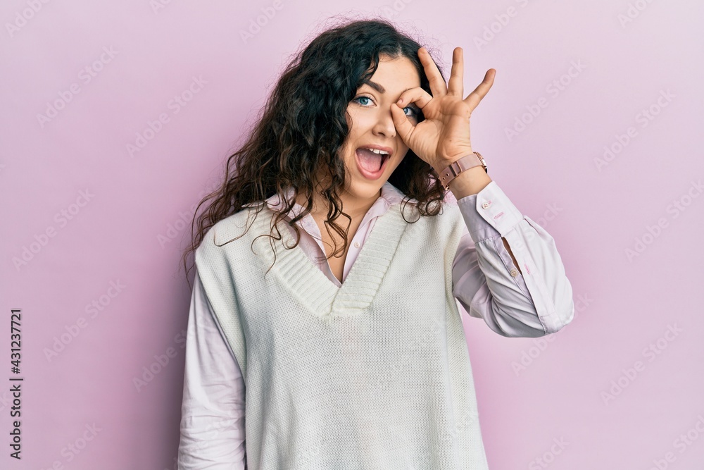 Young brunette woman with curly hair wearing casual clothes doing ok gesture with hand smiling, eye looking through fingers with happy face. Young brunette woman with curly hair wearing casual clothes doing ok gesture with hand smiling, eye looking through fingers with happy face.