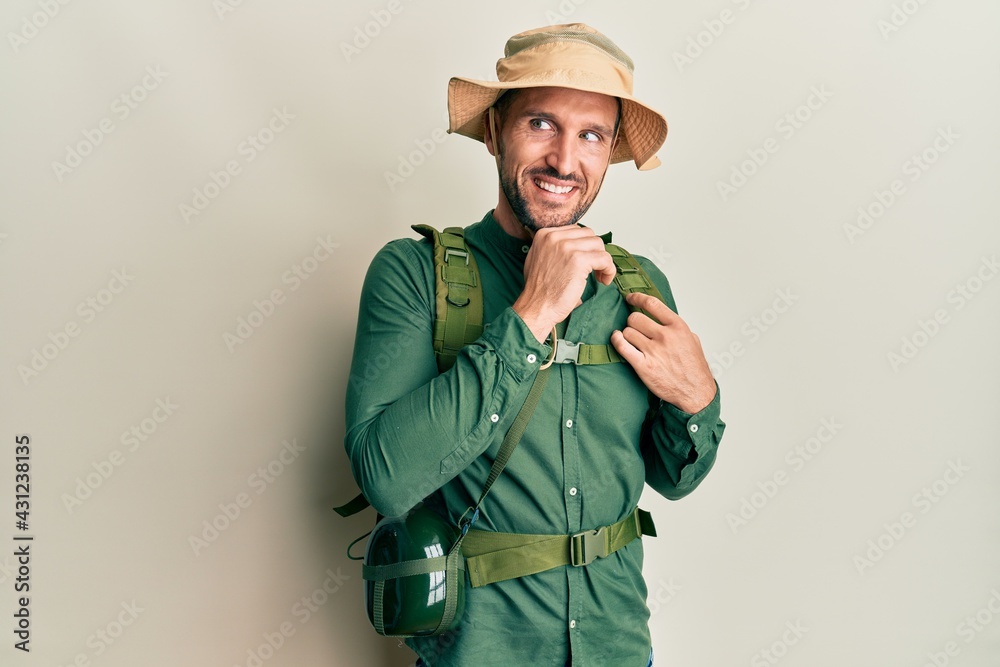 Handsome man with beard wearing explorer hat and backpack with hand on chin thinking about question, pensive expression. smiling and thoughtful face. doubt concept. Handsome man with beard wearing explorer hat and backpack with hand on chin thinking about question, pensive expression. smiling and thoughtful face. doubt concept.