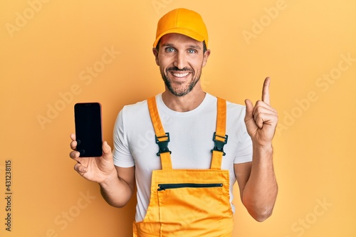 Young handsome man wearing handyman uniform showing smartphone screen smiling happy pointing with hand and finger to the side photo