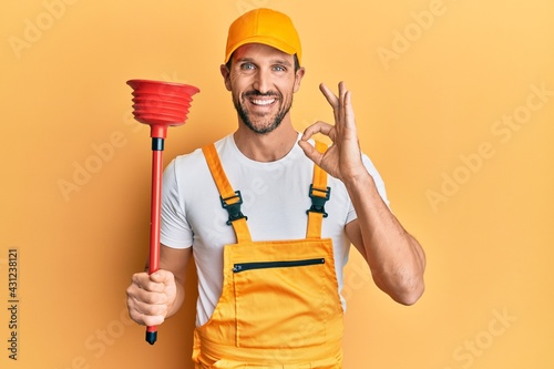 Фотография Young handsome man wearing plumber uniform holding toilet plunger doing ok sign