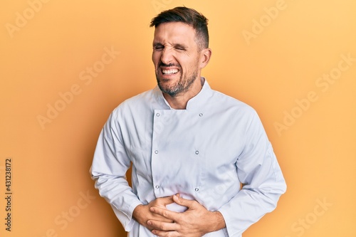 Handsome man with beard wearing professional cook uniform with hand on stomach because nausea, painful disease feeling unwell. ache concept.