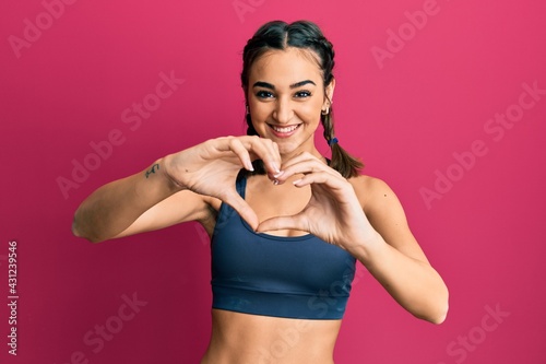 Fototapeta Naklejka Na Ścianę i Meble -  Young brunette girl wearing sportswear and braids smiling in love doing heart symbol shape with hands. romantic concept.
