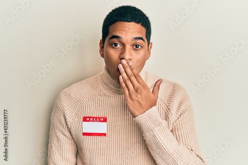 фотографія Young handsome hispanic man wearing hello my name is sticker identification covering mouth with hand, shocked and afraid for mistake