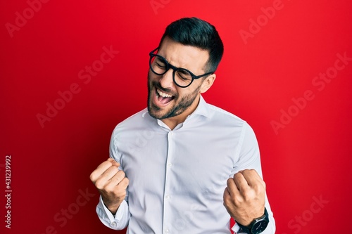 Young hispanic businessman wearing shirt and glasses very happy and excited doing winner gesture with arms raised, smiling and screaming for success. celebration concept.