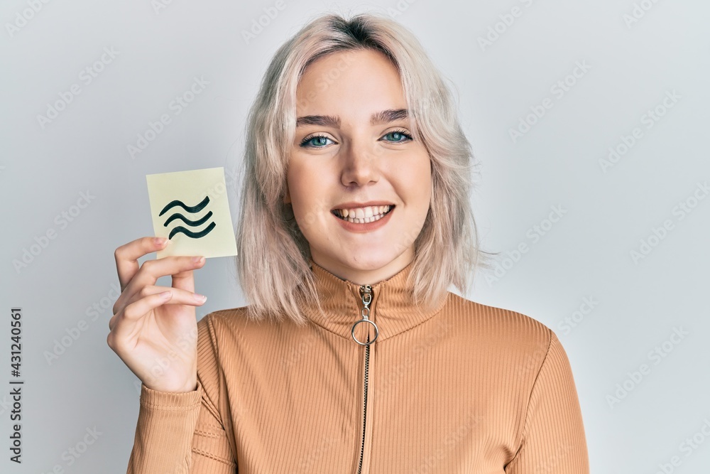 Young blonde girl holding paper with aquarius zodiac sign looking positive and happy standing and smiling with a confident smile showing teeth Young blonde girl holding paper with aquarius zodiac sign looking positive and happy standing and smiling with a confident smile showing teeth