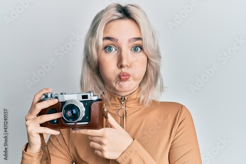 Young blonde girl holding vintage camera puffing cheeks with funny face. mouth inflated with air, catching air.