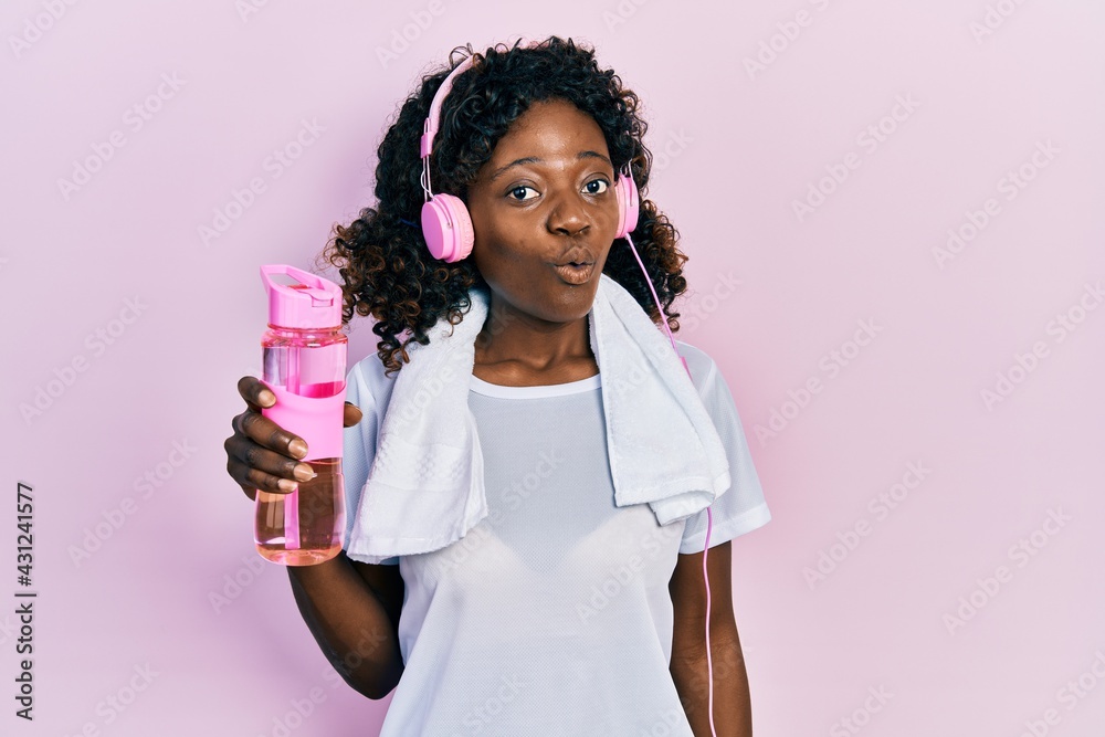 Young african american woman wearing sportswear holding water bottle using headphones scared and amazed with open mouth for surprise, disbelief face