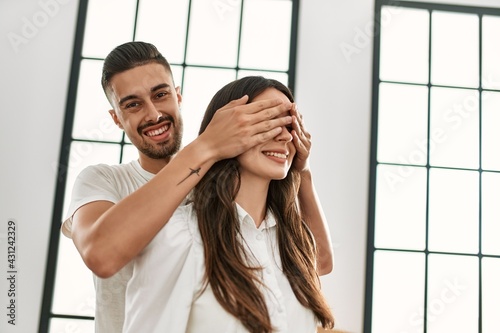Young hispanic man surprising woman putting hands on eyes at new home.