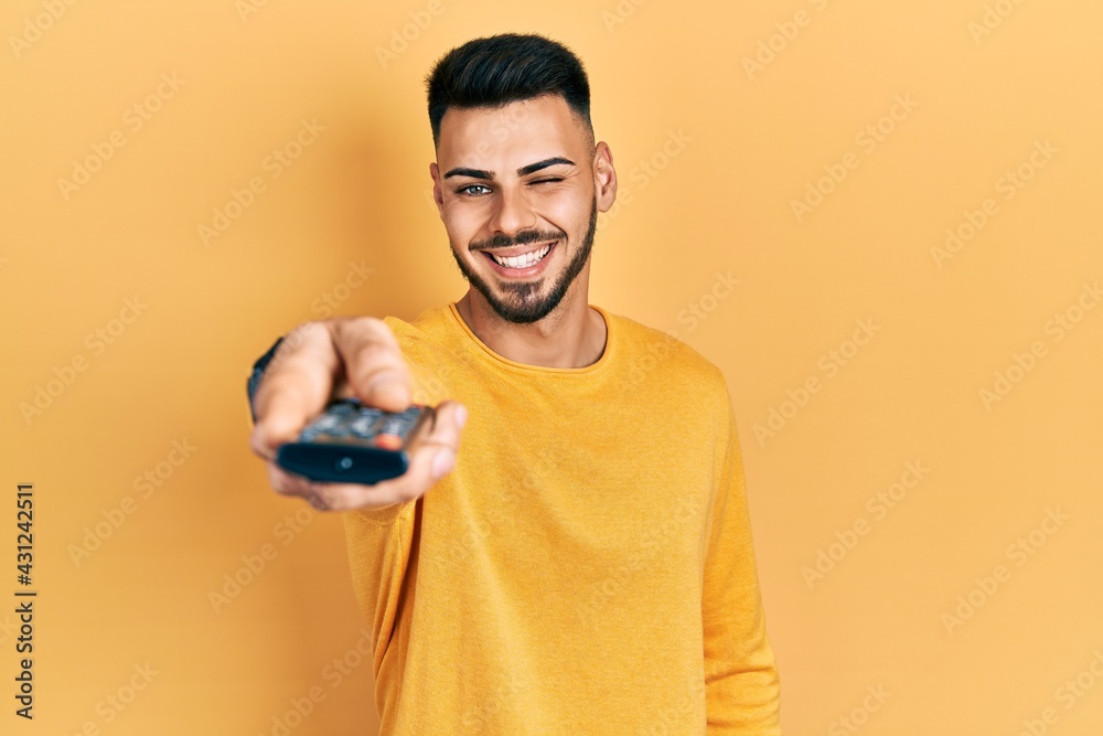 Young hispanic man with beard holding television remote control winking looking at the camera with sexy expression, cheerful and happy face. Young hispanic man with beard holding television remote control winking looking at the camera with sexy expression, cheerful and happy face.