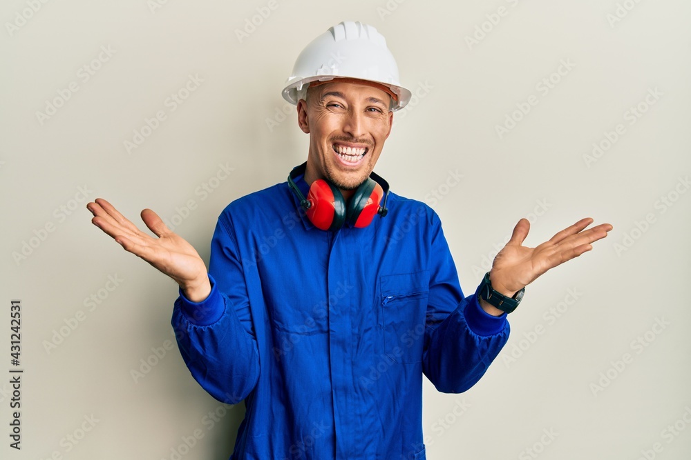 Bald man with beard wearing builder jumpsuit uniform and hardhat celebrating victory with happy smile and winner expression with raised hands