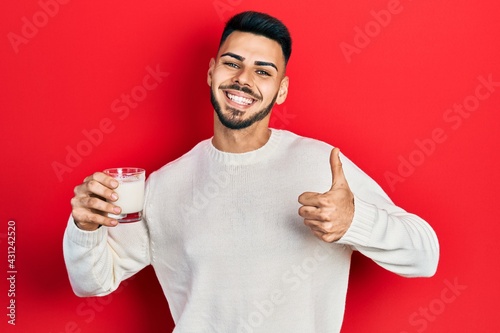 Young hispanic man with beard holding glass of milk smiling happy and positive, thumb up doing excellent and approval sign © Krakenimages.com