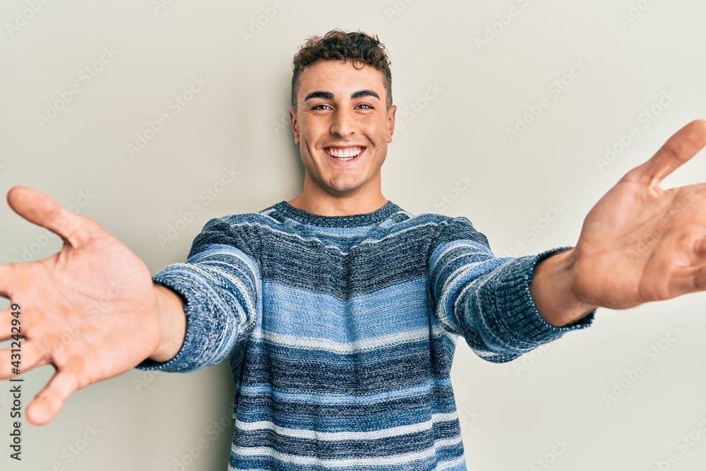 Hispanic young man wearing casual winter sweater looking at the camera smiling with open arms for hug. cheerful expression embracing happiness. Hispanic young man wearing casual winter sweater looking at the camera smiling with open arms for hug. cheerful expression embracing happiness.