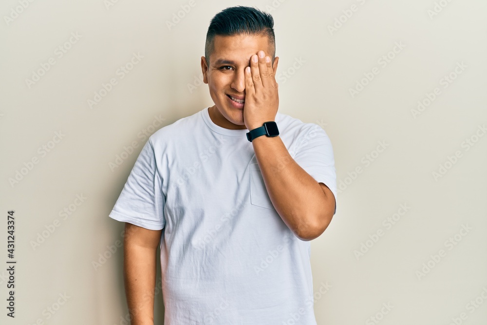 Young latin man wearing casual white t shirt covering one eye with hand, confident smile on face and surprise emotion. Young latin man wearing casual white t shirt covering one eye with hand, confident smile on face and surprise emotion.