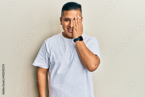 Young latin man wearing casual white t shirt covering one eye with hand, confident smile on face and surprise emotion.