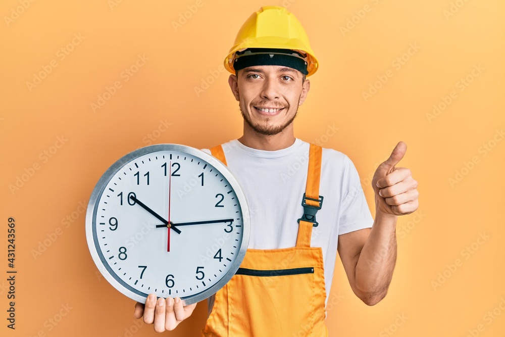Hispanic young man wearing builder uniform and hardhat holding clock smiling happy and positive, thumb up doing excellent and approval sign Hispanic young man wearing builder uniform and hardhat holding clock smiling happy and positive, thumb up doing excellent and approval sign