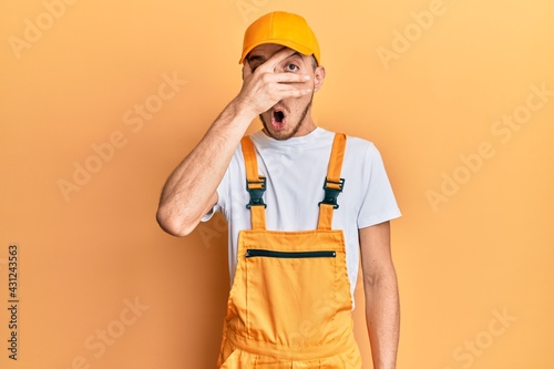 Hispanic young man wearing handyman uniform peeking in shock covering face and eyes with hand  looking through fingers with embarrassed expression.