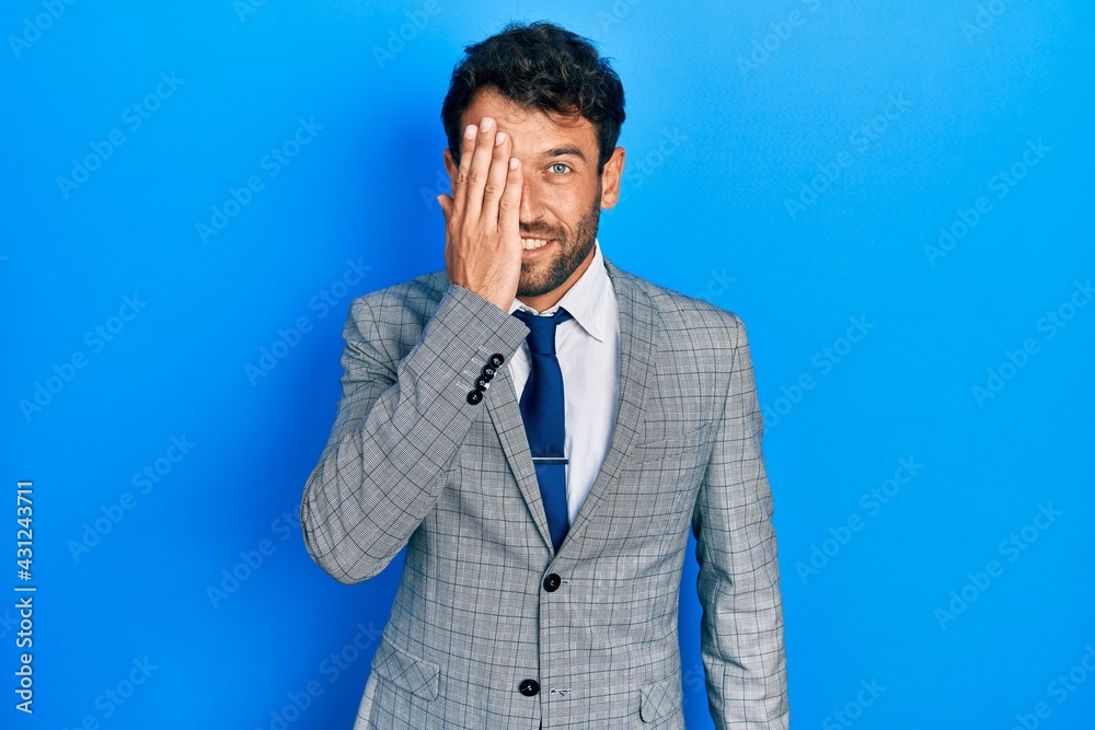 Handsome man with beard wearing business suit and tie covering one eye with hand, confident smile on face and surprise emotion. Handsome man with beard wearing business suit and tie covering one eye with hand, confident smile on face and surprise emotion.