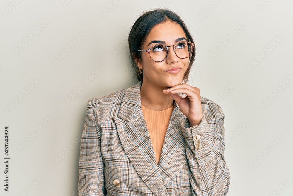 Young brunette woman wearing business jacket and glasses with hand on chin thinking about question, pensive expression. smiling and thoughtful face. doubt concept. Young brunette woman wearing business jacket and glasses with hand on chin thinking about question, pensive expression. smiling and thoughtful face. doubt concept.