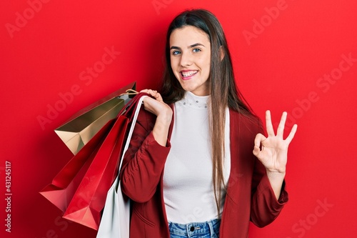 Young brunette teenager holding shopping bags doing ok sign with fingers, smiling friendly gesturing excellent symbol