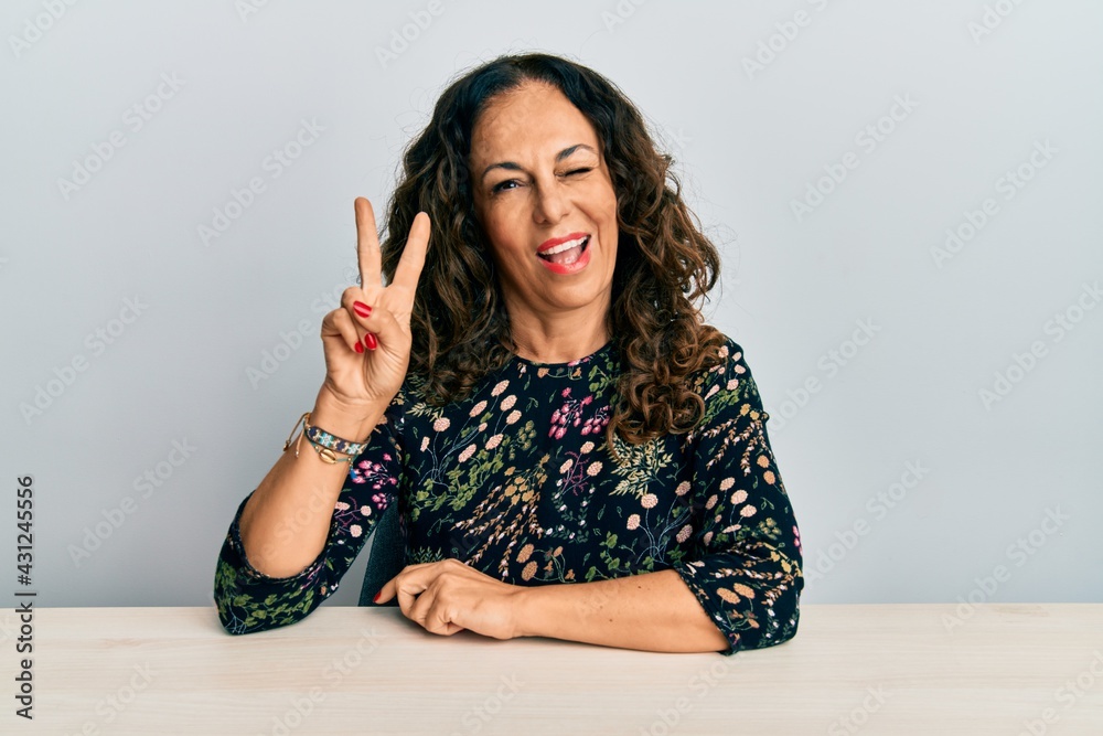 Beautiful middle age woman wearing casual clothes sitting on the table smiling with happy face winking at the camera doing victory sign. number two. Beautiful middle age woman wearing casual clothes sitting on the table smiling with happy face winking at the camera doing victory sign. number two.