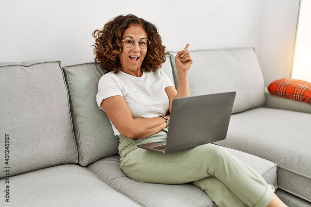 Middle age hispanic woman working using computer laptop at home smiling happy pointing with hand and finger to the side Middle age hispanic woman working using computer laptop at home smiling happy pointing with hand and finger to the side