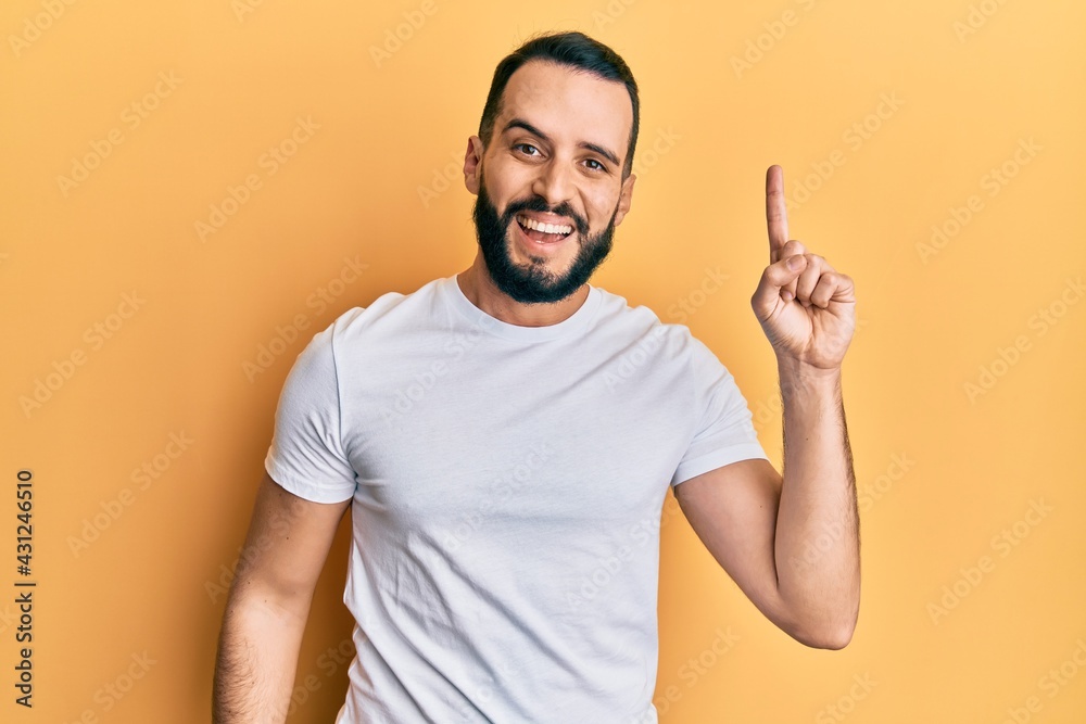 Young man with beard wearing casual white t shirt pointing finger up with successful idea. exited and happy. number one. Young man with beard wearing casual white t shirt pointing finger up with successful idea. exited and happy. number one.