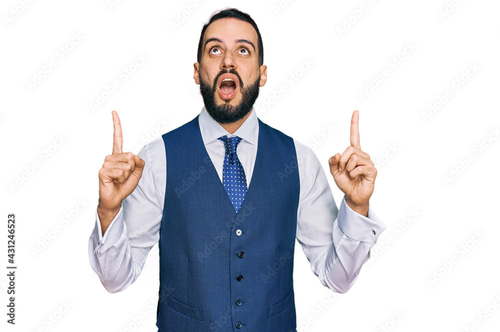 Young man with beard wearing business vest amazed and surprised looking up and pointing with fingers and raised arms. Young man with beard wearing business vest amazed and surprised looking up and pointing with fingers and raised arms.