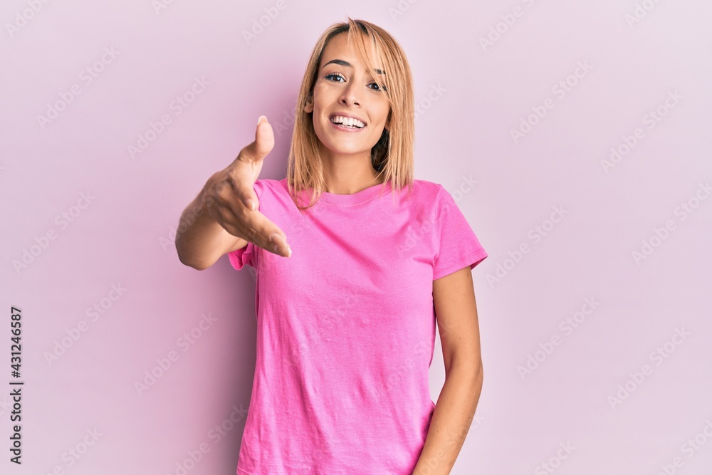 Beautiful blonde woman wearing casual pink tshirt smiling friendly offering handshake as greeting and welcoming. successful business. Beautiful blonde woman wearing casual pink tshirt smiling friendly offering handshake as greeting and welcoming. successful business.