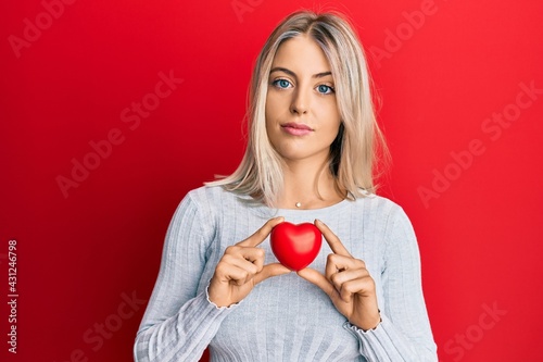 Beautiful blonde woman holding heart relaxed with serious expression on face. simple and natural looking at the camera.