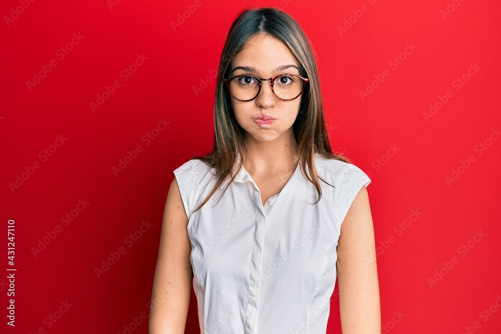 Young brunette woman wearing casual clothes and glasses puffing cheeks with funny face. mouth inflated with air, crazy expression. Young brunette woman wearing casual clothes and glasses puffing cheeks with funny face. mouth inflated with air, crazy expression.