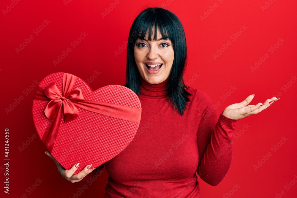 Young hispanic woman holding valentine gift celebrating achievement with happy smile and winner expression with raised hand Young hispanic woman holding valentine gift celebrating achievement with happy smile and winner expression with raised hand